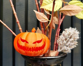Puede incluir: Una calabaza de Halloween naranja con una cara de miedo tallada en ella. La calabaza está en un jarrón negro con flores secas y ramas.