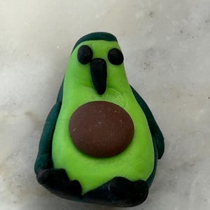 Könnte beinhalten: Eine grüne Figur in Form einer Avocado mit einem braunen Kern und schwarzen Augen, Schnabel und Füßen.