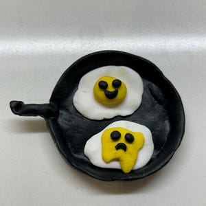 May include: A black frying pan with two fried eggs. One egg has a happy face and the other has a sad face.