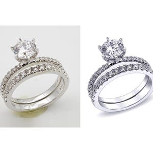 May include: A silver engagement ring set with a large, round, clear gemstone center stone. The band is encrusted with smaller, clear gemstones. The set includes a matching band with similar gemstone detailing.