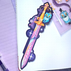 Pode incluir: Um autocolante colorido com uma espada de fantasia com uma empunhadura dourada e uma lâmina rosa e amarela. O autocolante tem um fundo roxo escuro com estrelas e um personagem de gato azul. Autocolantes adicionais são visíveis.