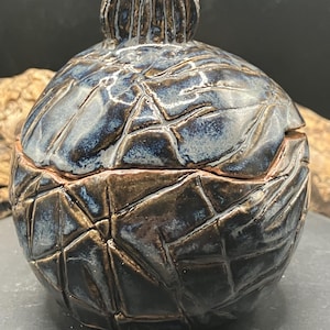 May include: A handmade ceramic jar with a lid. The jar is a dark blue and brown colour with a textured, geometric pattern. The lid has a knob on top. The jar is a decorative item.