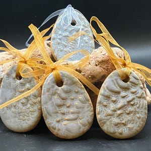 May include: Oval ceramic ornaments with textured surfaces and gold detailing. Each ornament has a hole at the top and is tied with a gold ribbon. The ornaments are displayed on rocks against a dark background.