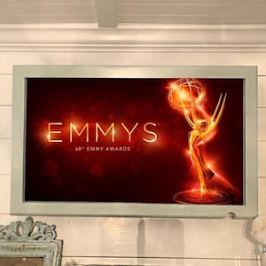 May include: A television screen displaying the 68th Emmy Awards logo. The logo features a gold statuette of a winged woman holding up a globe, with the word "EMMYS" in large, bold letters above the statuette. The background is a red gradient with a golden glow.