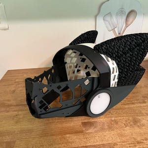 Op de afbeelding: Een 3D-geprint masker in dierstijl met een zwart geometrisch frame, een witte interne roosterstructuur en grote, getextureerde zwarte oren. De zijkant van het masker bevat een ronde witte schijf met de letters SL in het zwart gedrukt.