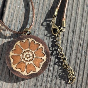 Puede incluir: Un colgante circular de madera con un diseño de flor de loto grabado a láser y un motivo central de ocho pétalos. El colgante está unido a un cordón marrón con un cierre de mosquetón de metal color bronce y una cadena ajustable.