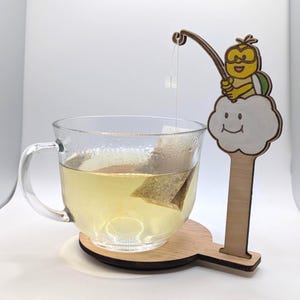 May include: A clear glass mug filled with tea, resting on a wooden coaster. A wooden tea bag holder shaped like a cartoon character fishing with a tea bag. The tea bag is suspended in the tea.