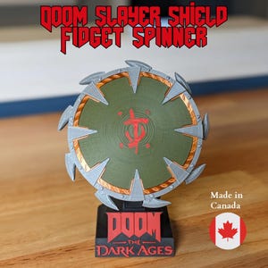 May include: A Doom Slayer Shield fidget spinner. The shield is olive green with a red symbol in the center and a silver and gold trim. The base reads "DOOM THE DARK AGES". The text "DOOM SLAYER SHIELD FIDGET SPINNER" is at the top.