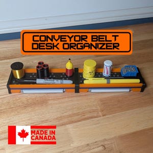 Puede incluir: Un organizador de escritorio negro y naranja con el texto "CONVEYOR BELT DESK ORGANIZER". El organizador contiene varios objetos en miniatura, incluyendo un bolígrafo, un rotulador y un pequeño barril. También se ven las palabras "MADE IN CANADA".