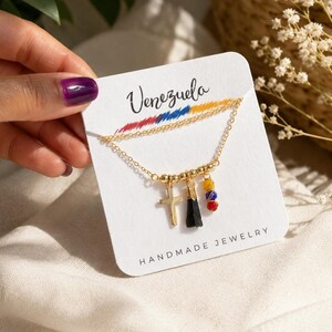 Op de afbeelding: Een goudkleurige ketting op een witte kaart met de tekst Venezuela en Handmade Jewelry. De ketting heeft drie bedels: een gouden kruis, een zwarte stenen hanger en een klein groepje van gele, blauwe en rode kralen aan een gouden draad.