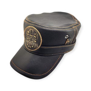 May include: Black leather military-style cap with a gold skull emblem on the front. The cap features a short brim, gold stitching, and a strap detail. The cap is a dark color and has a textured appearance.