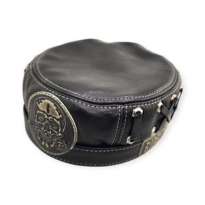 May include: Black leather hat with a round crown and a decorative skull emblem. The hat features white stitching and metal accents. A patch with the word "Riders" is visible.