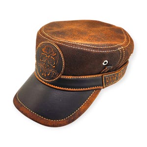 May include: Brown leather cap with a black brim and a skull emblem. The cap features white stitching and a band that reads "RIDE TO". The cap is a military style hat.