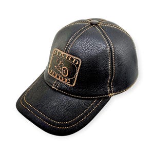 Può includere: Cappellino da baseball in pelle nera con visiera curva e cuciture. Il cappellino presenta una toppa rettangolare con le parole "HARD RIDE" e una grafica di una motocicletta. Le cuciture e la toppa sono di colore marrone.