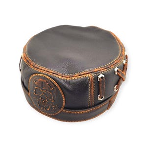 May include: A black leather cap with a skull design. The cap features brown stitching and accents, with a skull emblem on the side. The cap has a round shape and is made of leather.