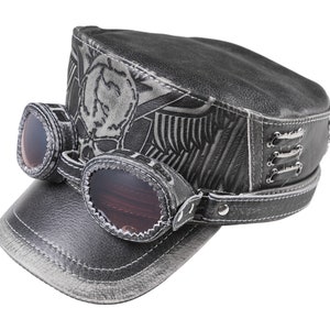 May include: A gray leather cap with a skull and wings design. The cap has a brim and a strap with a buckle. The cap also has goggles attached to the front.