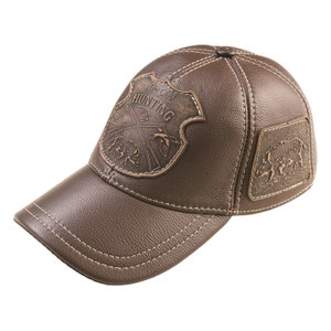 May include: A brown leather baseball cap with a stitched brim. The cap features a brown leather patch with the text "The Club Hunting" and a design of crossed rifles and a hunting scene. There is a smaller brown leather patch on the side of the cap with a boar design.
