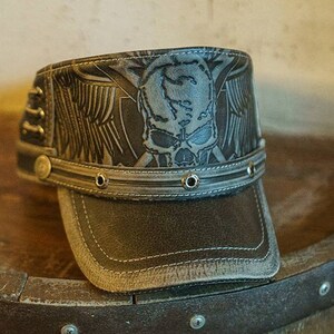 May include: A brown leather baseball cap with a skull and wings design. The cap has a gray brim and a black leather strap with metal rivets.