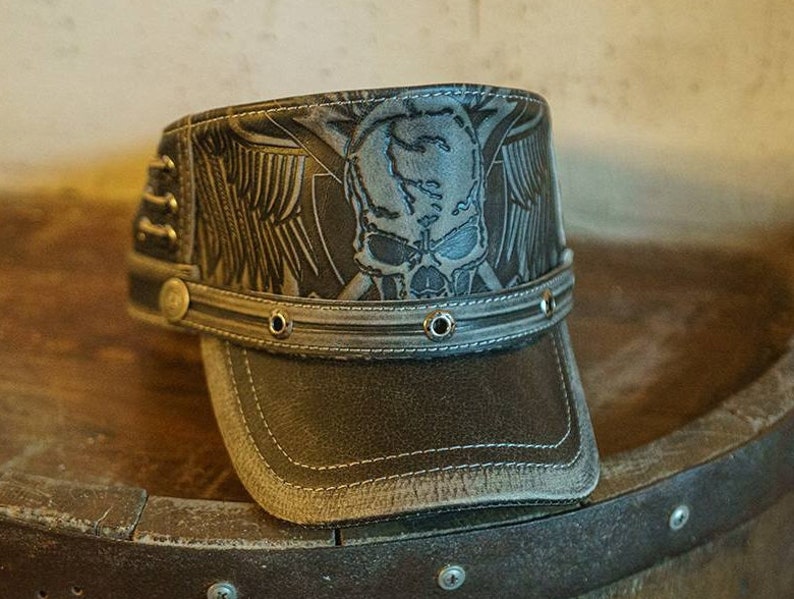May include: A brown leather baseball cap with a skull and wings design. The cap has a gray brim and a black leather strap with metal rivets.