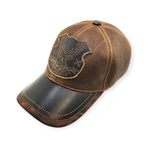 May include: Brown leather baseball cap with a black bill. The cap features a decorative emblem with a deer head and foliage. The cap is stitched with a contrasting thread and has a button on top.