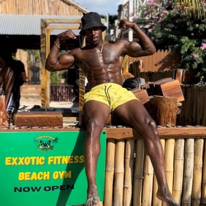 Puede incluir: Un hombre sin camisa que lleva un sombrero negro y pantalones de baño amarillos posa con los brazos en alto. Está de pie frente a una valla de madera con un cartel que dice "Exxotic Fitness Beach Gym Now Open".