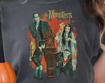 Peut inclure: Un t-shirt gris foncé avec une illustration vintage de la famille Munster. L'illustration représente les personnages de la série télévisée classique, dont Herman Munster, Lily Munster, Grandpa Munster, Eddie Munster et Marilyn Munster. Le texte "The Munsters" est imprimé en blanc au-dessus de l'illustration.