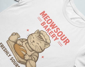 Könnte beinhalten: Weißes T-Shirt mit einem Cartoon-Katzenbäcker, der eine Kochmütze und eine Schürze trägt und Brot hält. Der Text "MEOWSOUR BAKERY" und "FRESHLY SOURDOUGH" ist rot. Das Design umfasst eine Kanne und eine Schüssel.