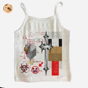 May include: Cream camisole top with a graphic design featuring a silver cross, Japanese text, and other symbols. The top has thin straps. The design includes a biohazard symbol and a smiling face. The top is made of a soft material.