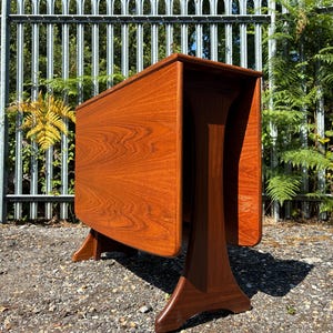 May include: A wooden drop-leaf table with a warm brown finish and visible wood grain patterns. The table features a rectangular top with two hinged side leaves currently in a folded position, supported by a central pedestal base with curved legs.