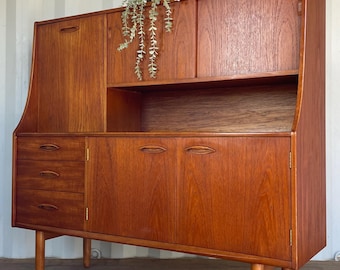 May include: A mid-century modern wooden cabinet with a warm, reddish-brown finish. It features multiple storage compartments, including drawers and doors, and is raised on tapered legs. A small plant sits atop the cabinet.