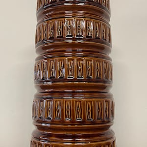 May include: A tall, brown ceramic vase with a glossy finish. The vase has a cylindrical shape with a series of stacked, textured bands. Each band features a repeating vertical pattern. The vase is a decorative item.