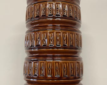 May include: A tall, brown ceramic vase with a glossy finish. The vase has a cylindrical shape with a series of stacked, textured bands. Each band features a repeating vertical pattern. The vase is a decorative item.