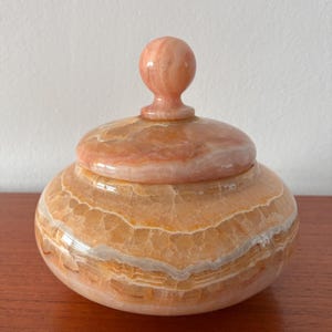 May include: A polished, round stone jar with a matching lid featuring a spherical knob handle. The vessel displays natural horizontal banding in shades of translucent peach, warm orange, and creamy white, showcasing a smooth, glossy finish.