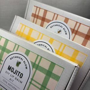 May include: Three silver boxes with green, yellow, and brown plaid patterns. Each box has a circular sticker with "Botanical Glow." The front box reads "Mojito DIY Lip Balm Kit 100% Natural."