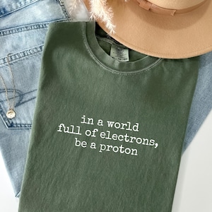 May include: An olive green t-shirt with the text "in a world full of electrons, be a proton" in white. The shirt is paired with light wash denim jeans, a gold necklace, and a tan hat. Dried flowers are also present.