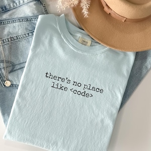 May include: A light blue t-shirt with the text "there's no place like <code >" in black. The shirt is paired with light wash denim jeans, a tan hat, and a gold necklace. The word "chambray" is printed at the bottom.