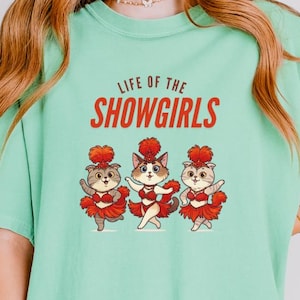 May include: Mint green t-shirt with the text "LIFE OF THE SHOWGIRLS" in red. The graphic features three cartoon cats in red and white outfits. The cats are wearing red headpieces and are dancing.