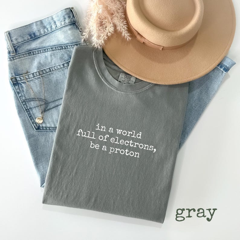 May include: A gray t-shirt with the text "in a world full of electrons, be a proton" in white. The shirt is paired with light blue jeans, a tan hat, and a gold necklace.