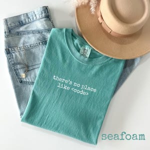 May include: A seafoam green t-shirt with the text "there's no place like <code >" in white, paired with light wash denim jeans, a tan hat, and a gold necklace. The word "seafoam" is printed at the bottom.