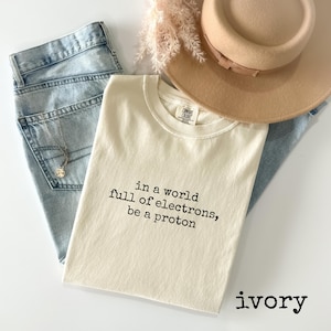 May include: An ivory-colored t-shirt with the text "in a world full of electrons, be a proton" in black. The shirt is folded and placed on top of light blue jeans, a beige hat, and a necklace. The word "ivory" is printed at the bottom.