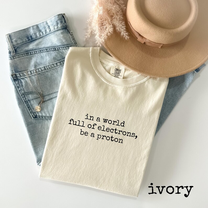 May include: An ivory-colored t-shirt with the text "in a world full of electrons, be a proton" in black. The shirt is folded and placed on top of light blue jeans, a beige hat, and a necklace. The word "ivory" is printed at the bottom.