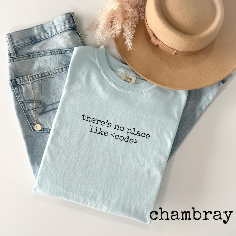 May include: A light blue t-shirt with the text "there's no place like <code >" in black. The shirt is paired with light wash denim jeans, a tan hat, and a gold necklace. The word "chambray" is printed at the bottom.