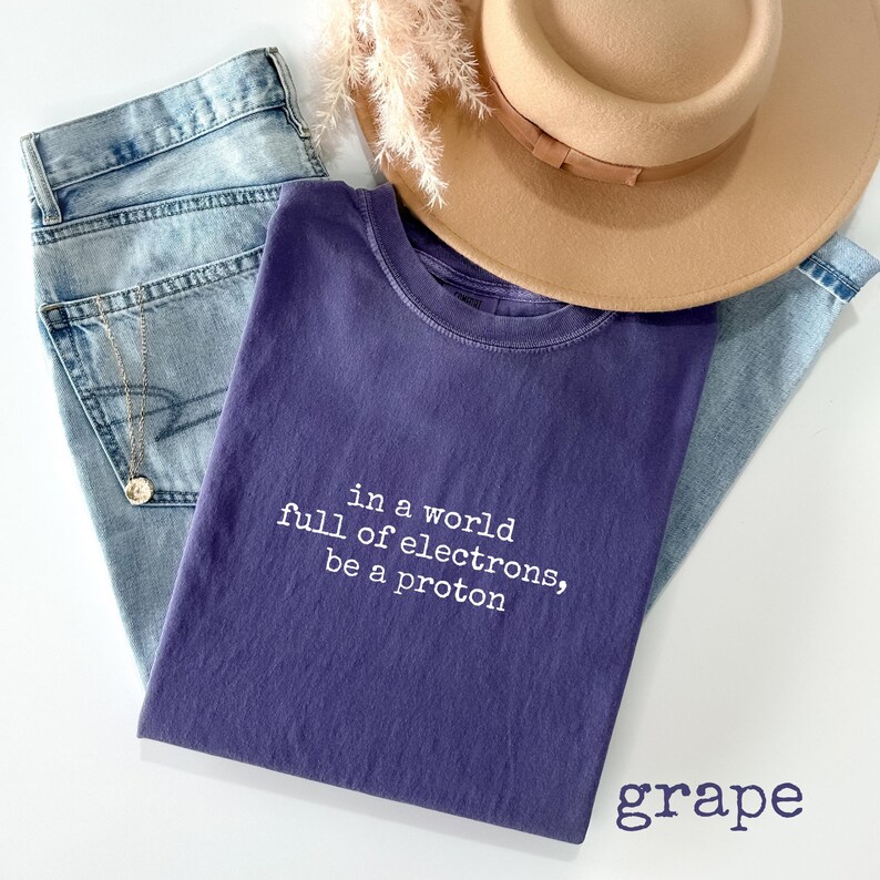 May include: A grape-colored t-shirt with the text "in a world full of electrons, be a proton" in white. The shirt is paired with light blue jeans, a tan hat, and a gold necklace. The word "grape" is printed at the bottom right.