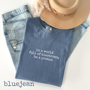 May include: A blue t-shirt with the text "in a world full of electrons, be a proton" is laid on top of a pair of blue jeans. A tan hat and a gold necklace are also in the image. The word "bluejean" is at the bottom.