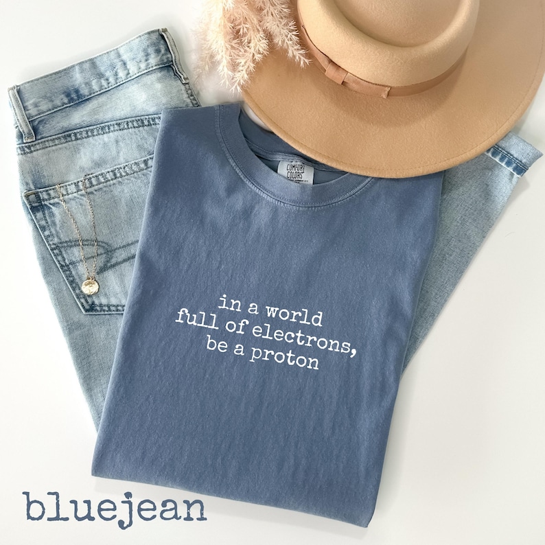 May include: A blue t-shirt with the text "in a world full of electrons, be a proton" is laid on top of a pair of blue jeans. A tan hat and a gold necklace are also in the image. The word "bluejean" is at the bottom.