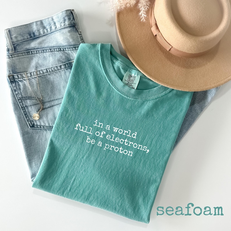 May include: A seafoam green t-shirt with the text "in a world full of electrons, be a proton" in white. The shirt is paired with light wash denim jeans, a gold necklace, and a tan fedora hat. The word "seafoam" is printed at the bottom.