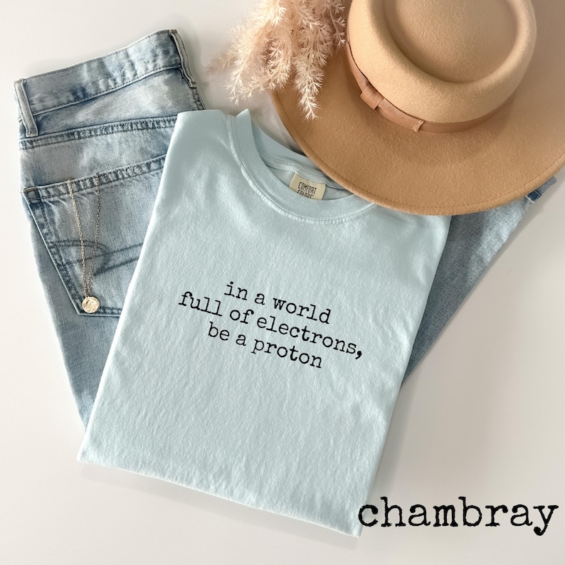 May include: A light blue t-shirt with the text "in a world full of electrons, be a proton" in black. The shirt is paired with light wash denim jeans and a tan hat. Dried flowers are also in the image.