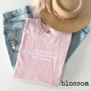 May include: A light pink t-shirt with the text "in a world full of electrons, be a proton" in white. The shirt is paired with light wash denim jeans, a tan hat, and a gold necklace.