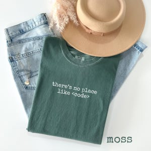 May include: A casual fashion flat lay featuring a moss green t-shirt with the text "there's no place like <code">, light wash denim jeans, a tan fedora hat, and a gold necklace. The word "moss" is printed in the bottom right corner.