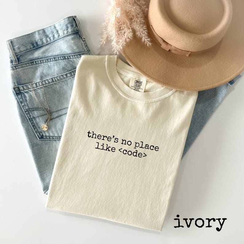 May include: Ivory-colored t-shirt with the text "there's no place like <code >" in black. The shirt is folded and placed with light blue jeans, a tan hat, and a necklace. Dried flowers are also in the image.